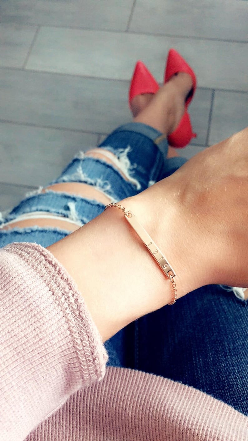 May include: A gold chain bracelet with a thin bar charm engraved with the numbers "1319". The bracelet is worn on a person's wrist.