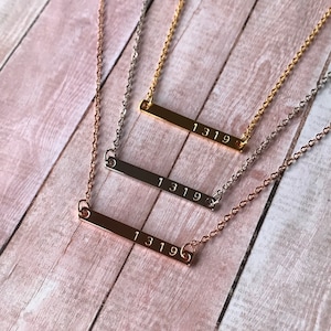 May include: Three gold bar necklaces with the number 1319 engraved on each. The necklaces are layered on a wooden surface.