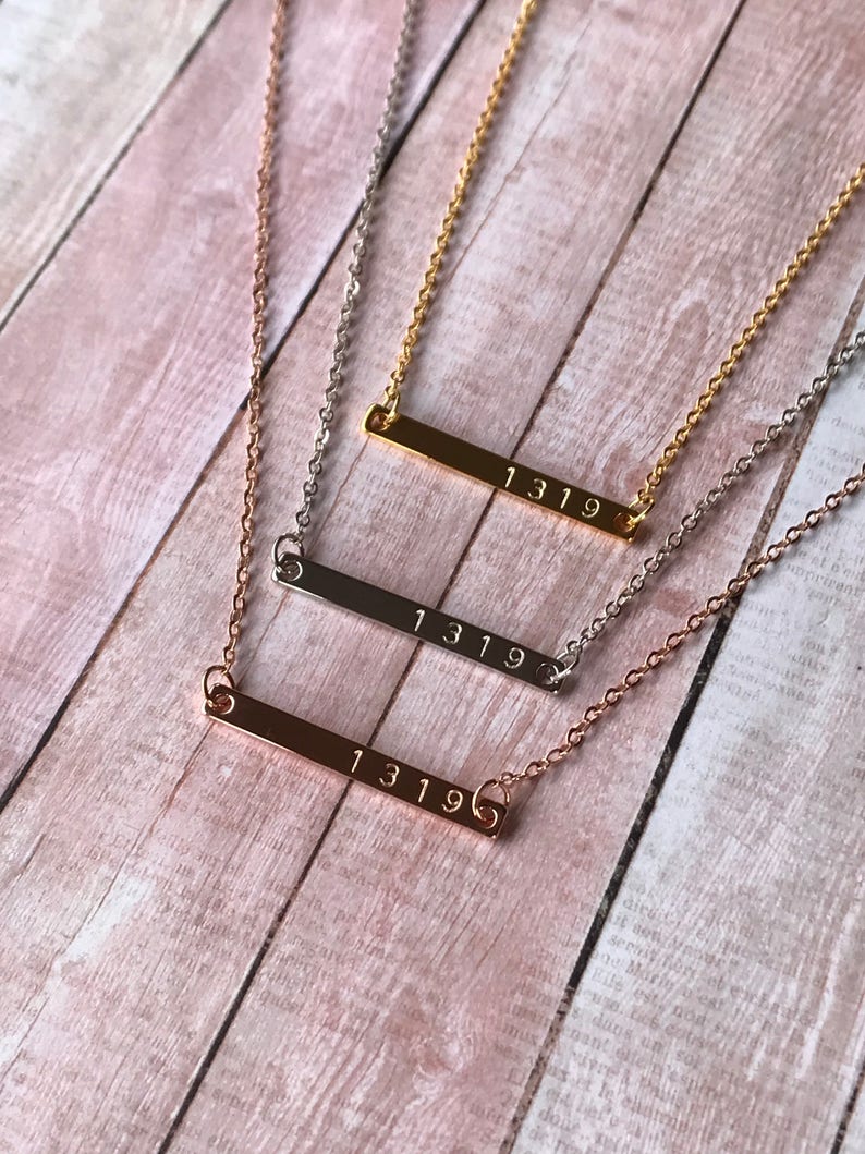 May include: Three gold bar necklaces with the number 1319 engraved on each. The necklaces are layered on a wooden surface.