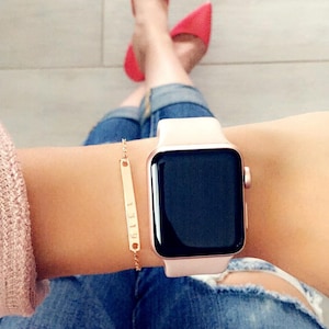 May include: A rose gold Apple Watch with a white band on a person's wrist. The watch has a black face and a small gold bracelet with a bar charm is on the same wrist.