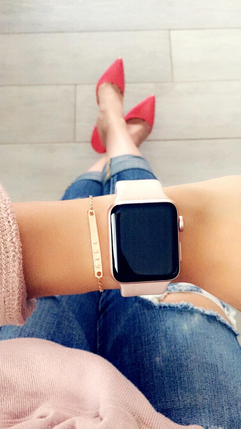 May include: A rose gold Apple Watch with a white band on a person's wrist. The watch has a black face and a small gold bracelet with a bar charm is on the same wrist.