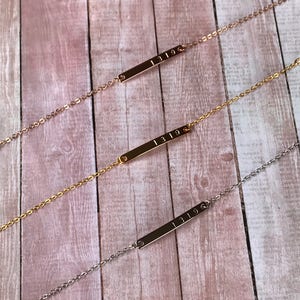 May include: Three gold bar necklaces with the number "1319" engraved on each. The necklaces are on a wooden background.