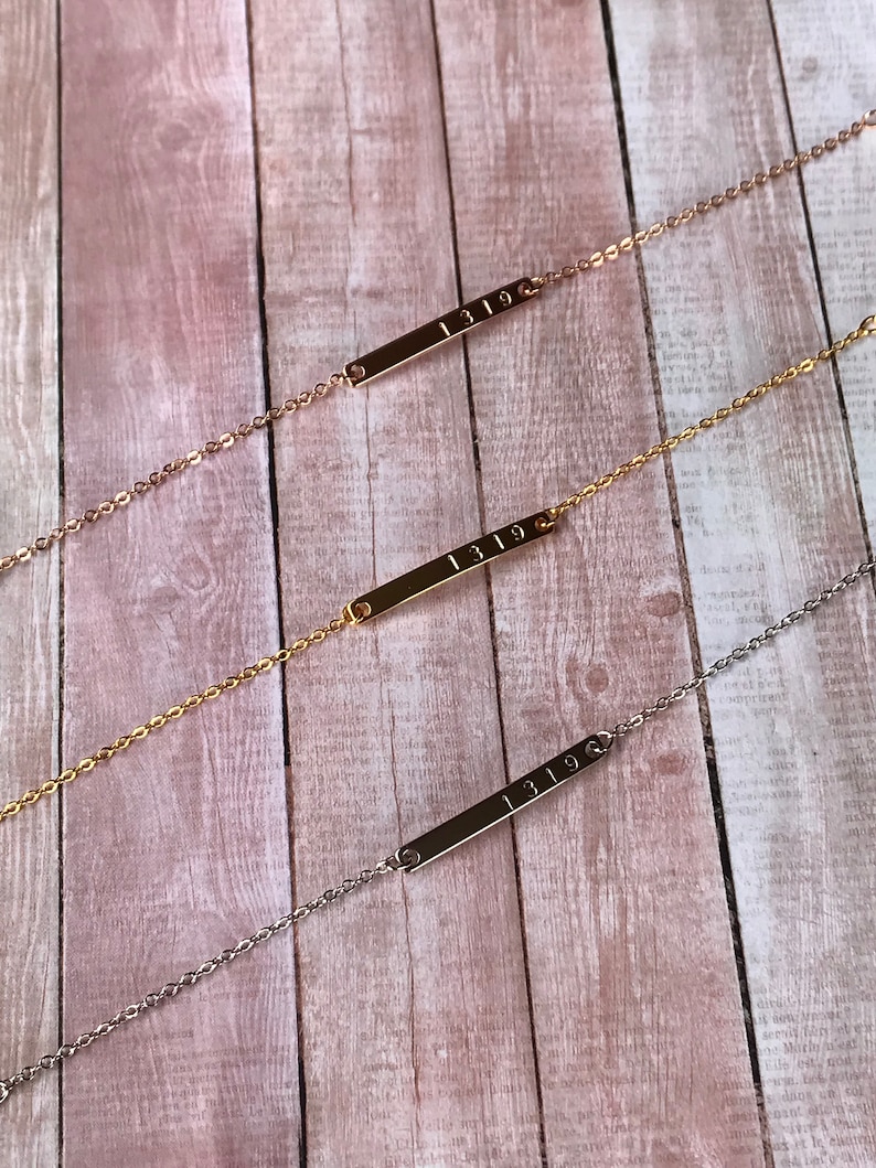May include: Three gold bar necklaces with the number "1319" engraved on each. The necklaces are on a wooden background.