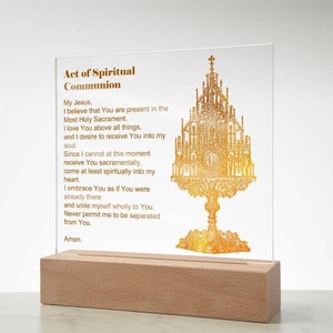 May include: A clear acrylic plaque with the text "Act of Spiritual Communion" and a gold-colored ornate religious illustration. The plaque is set on a light-colored wooden base.