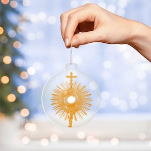 Puede incluir: Un adorno de Navidad de vidrio transparente con una imagen dorada de un copón, un recipiente sagrado utilizado en el culto católico, en su interior. El adorno está colgado de una cuerda plateada.