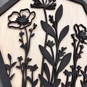 Puede incluir: Arte de pared de madera con un dise&ntilde;o floral negro sobre un fondo de madera clara. El dise&ntilde;o incluye varias flores y follaje, todos recortados en silueta. El marco tiene una forma geom&eacute;trica oscura, a&ntilde;adiendo un toque moderno.