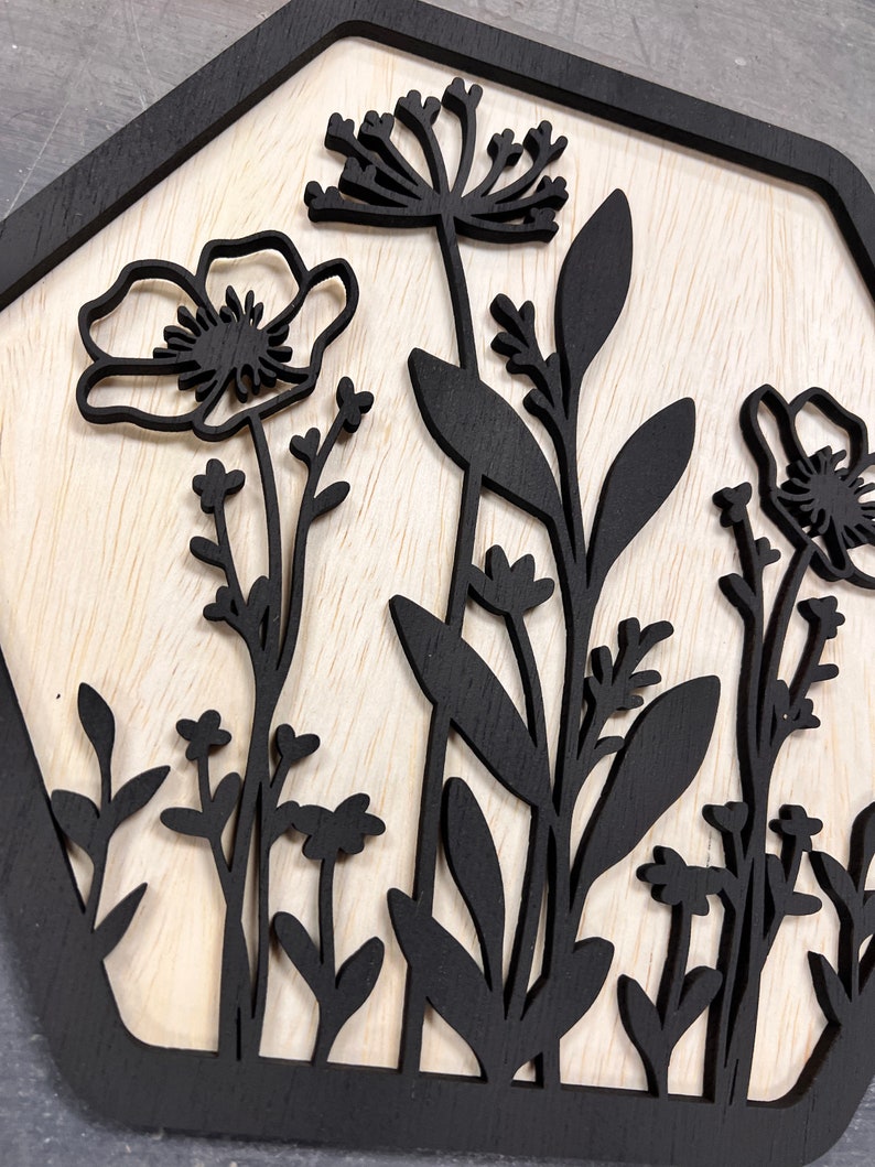 Puede incluir: Arte de pared de madera con un dise&ntilde;o floral negro sobre un fondo de madera clara. El dise&ntilde;o incluye varias flores y follaje, todos recortados en silueta. El marco tiene una forma geom&eacute;trica oscura, a&ntilde;adiendo un toque moderno.