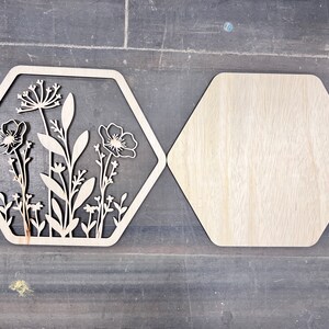 Puede incluir: Dos piezas decorativas de madera con forma de hex&aacute;gono. Una presenta un dise&ntilde;o floral cortado con l&aacute;ser con flores y hojas. La otra es un hex&aacute;gono de madera liso. Las piezas son de color marr&oacute;n claro y est&aacute;n sobre una superficie gris oscuro.