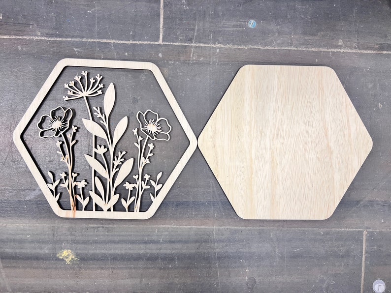 Puede incluir: Dos piezas decorativas de madera con forma de hex&aacute;gono. Una presenta un dise&ntilde;o floral cortado con l&aacute;ser con flores y hojas. La otra es un hex&aacute;gono de madera liso. Las piezas son de color marr&oacute;n claro y est&aacute;n sobre una superficie gris oscuro.