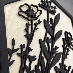 Puede incluir: Arte de pared de madera con un dise&ntilde;o floral negro sobre un fondo de madera clara. Las flores y el follaje negros est&aacute;n cortados con l&aacute;ser, creando un efecto de capas dentro de un marco hexagonal negro. La obra de arte tiene una est&eacute;tica natural y r&uacute;stica.