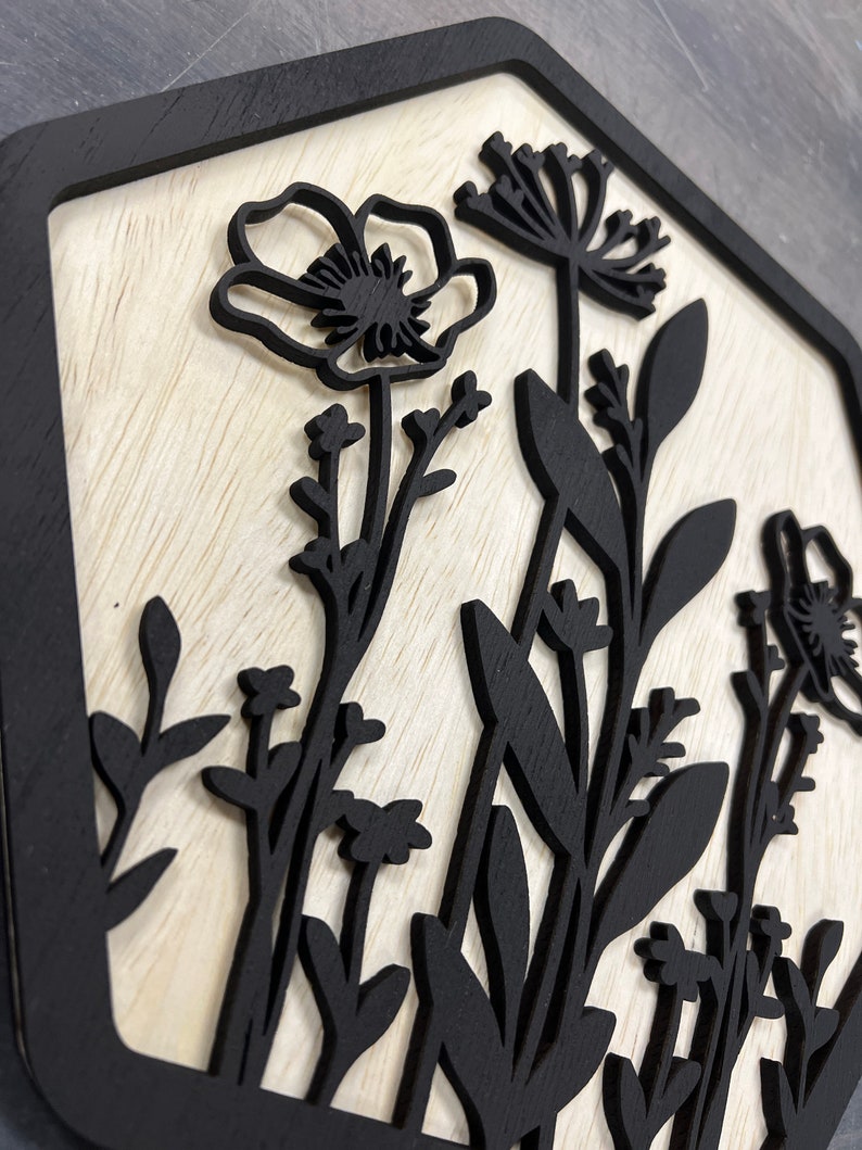 Puede incluir: Arte de pared de madera con un dise&ntilde;o floral negro sobre un fondo de madera clara. Las flores y el follaje negros est&aacute;n cortados con l&aacute;ser, creando un efecto de capas dentro de un marco hexagonal negro. La obra de arte tiene una est&eacute;tica natural y r&uacute;stica.
