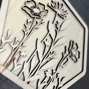 Puede incluir: Un dise&ntilde;o floral de madera cortado con l&aacute;ser en un marco hexagonal. La obra de arte presenta contornos detallados de varias flores y follaje, con un acabado de veta de madera natural. El dise&ntilde;o est&aacute; en capas, creando un efecto tridimensional.