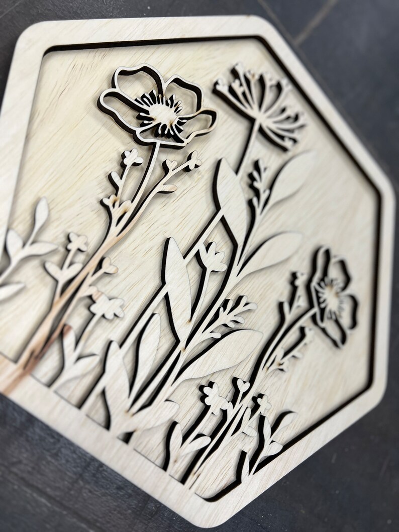 Puede incluir: Un dise&ntilde;o floral de madera cortado con l&aacute;ser en un marco hexagonal. La obra de arte presenta contornos detallados de varias flores y follaje, con un acabado de veta de madera natural. El dise&ntilde;o est&aacute; en capas, creando un efecto tridimensional.
