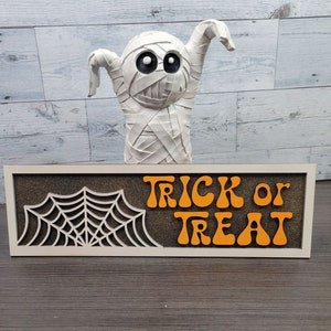 Könnte beinhalten: Ein weißes Holzschild mit einem schwarzen Spinnennetz-Ausschnitt und den Worten "Trick or Treat" in orangefarbenen Buchstaben.