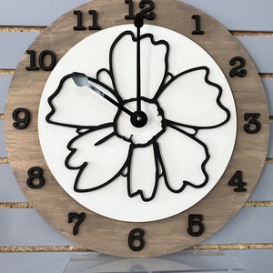 May include: A wooden clock with a white flower design on the face. The clock has black numbers and hands. The clock is on a clear stand.