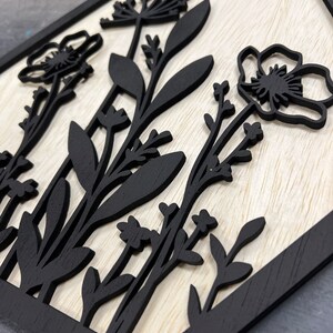 Puede incluir: Arte de pared de madera con un dise&ntilde;o floral negro sobre un fondo claro de vetas de madera. La obra de arte tiene una forma geom&eacute;trica con un borde negro. Los elementos florales incluyen flores y hojas estilizadas.