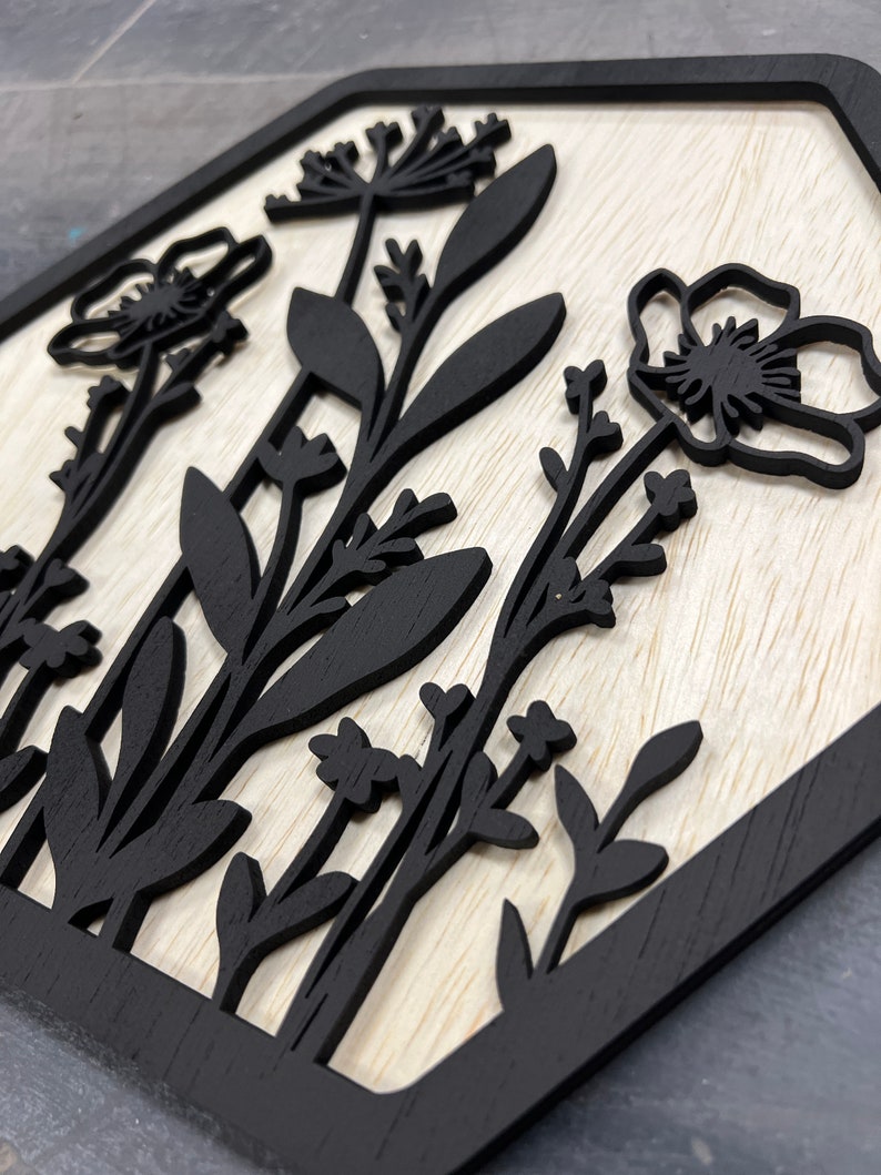 Puede incluir: Arte de pared de madera con un dise&ntilde;o floral negro sobre un fondo claro de vetas de madera. La obra de arte tiene una forma geom&eacute;trica con un borde negro. Los elementos florales incluyen flores y hojas estilizadas.