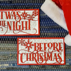 Peut inclure: Deux panneaux en bois rouge avec du texte blanc et des flocons de neige. Le panneau du haut indique "Twas the Night" et le panneau du bas indique "Before Christmas".