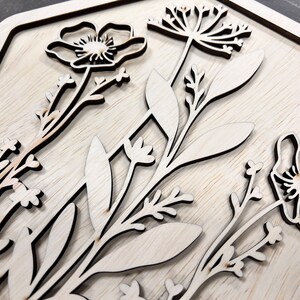 Puede incluir: Obra de arte floral de madera con flores y hojas cortadas con l&aacute;ser. El dise&ntilde;o incluye varios elementos bot&aacute;nicos, como amapolas y delicadas ramitas, dispuestos dentro de un marco cuadrado. La veta natural de la madera a&ntilde;ade un toque r&uacute;stico.