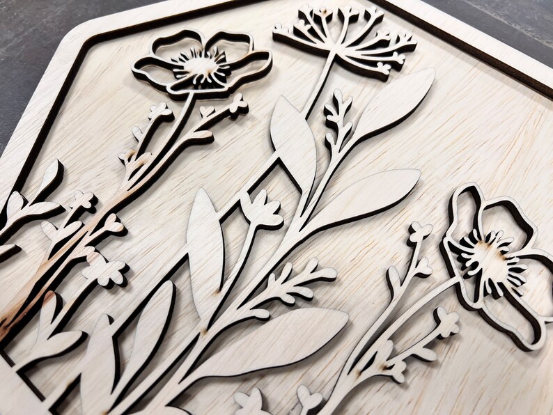Puede incluir: Obra de arte floral de madera con flores y hojas cortadas con l&aacute;ser. El dise&ntilde;o incluye varios elementos bot&aacute;nicos, como amapolas y delicadas ramitas, dispuestos dentro de un marco cuadrado. La veta natural de la madera a&ntilde;ade un toque r&uacute;stico.