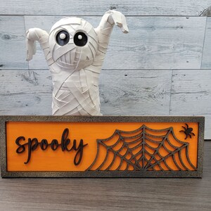 Könnte beinhalten: Ein Holzschild mit orangefarbenem Hintergrund und schwarzer Aufschrift "spooky". Rechts auf dem Schild befindet sich ein schwarzes Spinnennetz mit einer Spinne. Hinter dem Schild steht eine weiße Mumienfigur.