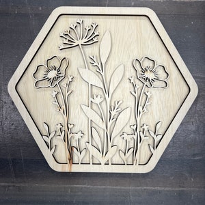 Puede incluir: Arte mural floral de madera. La obra presenta un dise&ntilde;o cortado con l&aacute;ser de varias flores y follaje dentro de un marco hexagonal. La veta natural de la madera proporciona una paleta de colores neutros, adecuada para la decoraci&oacute;n del hogar.