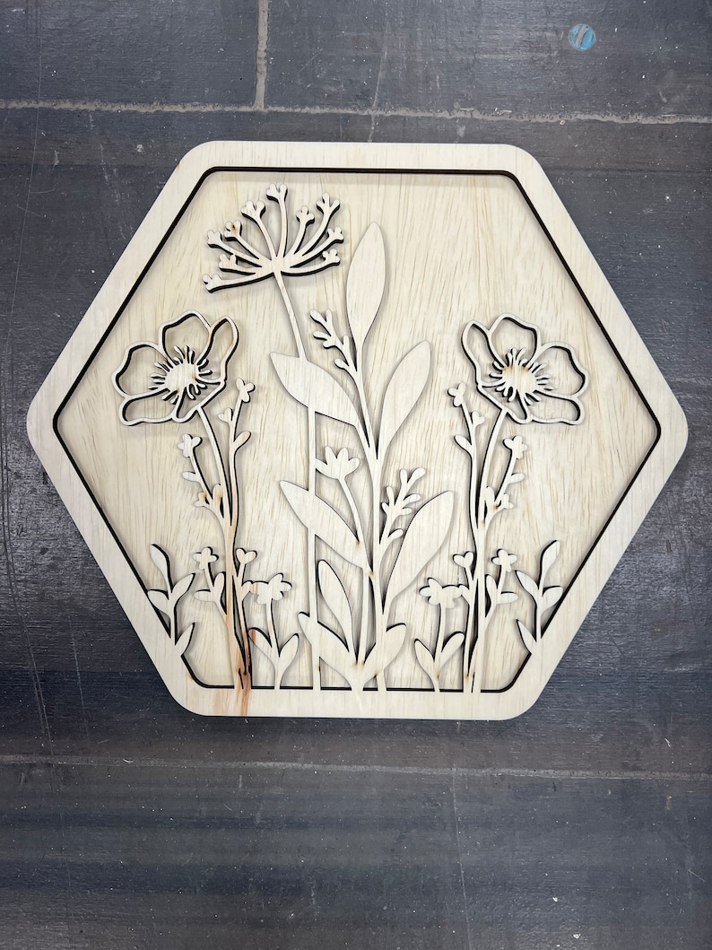 Puede incluir: Arte mural floral de madera. La obra presenta un dise&ntilde;o cortado con l&aacute;ser de varias flores y follaje dentro de un marco hexagonal. La veta natural de la madera proporciona una paleta de colores neutros, adecuada para la decoraci&oacute;n del hogar.