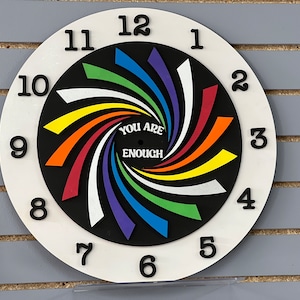 Puede incluir: Una esfera de reloj blanca con números negros del 1 al 12. La esfera del reloj tiene un centro negro con un diseño de remolino arcoíris y las palabras "You Are Enough".