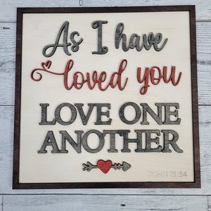 Op de afbeelding: Een houten bord met de tekst "As I have loved you, love one another" in grijze en rode letters. Onder de tekst staat een klein rood hart met een pijl. De tekst "JOHN 13:34" staat in de rechterbenedenhoek.