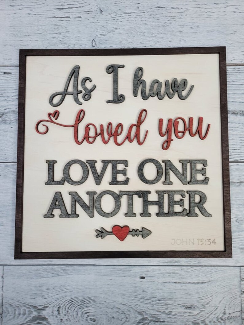 Op de afbeelding: Een houten bord met de tekst "As I have loved you, love one another" in grijze en rode letters. Onder de tekst staat een klein rood hart met een pijl. De tekst "JOHN 13:34" staat in de rechterbenedenhoek.