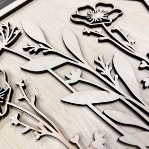 Puede incluir: Arte mural floral de madera con flores y hojas cortadas con l&aacute;ser. El dise&ntilde;o incluye amapolas y otras flores silvestres, con un acabado natural de vetas de madera. La obra de arte est&aacute; montada sobre un fondo de madera clara.