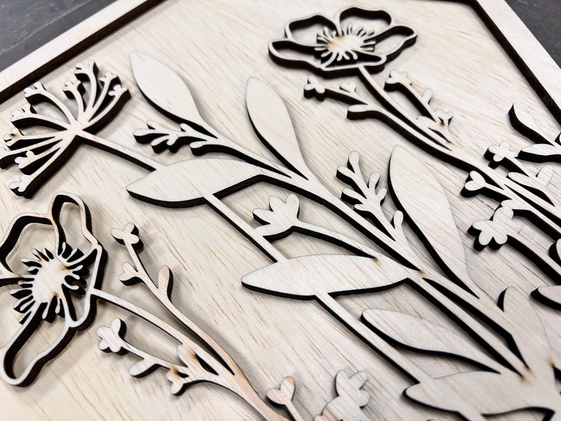 Puede incluir: Arte mural floral de madera con flores y hojas cortadas con l&aacute;ser. El dise&ntilde;o incluye amapolas y otras flores silvestres, con un acabado natural de vetas de madera. La obra de arte est&aacute; montada sobre un fondo de madera clara.