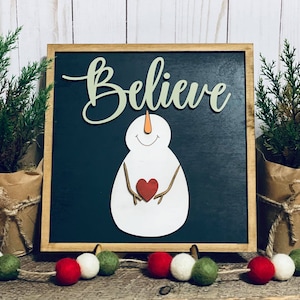 May include: A wooden sign with a black chalkboard background featuring a white snowman holding a red heart. The sign says "Believe" in green cursive lettering.