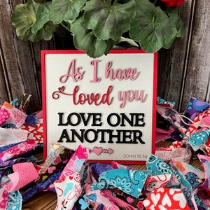 Op de afbeelding: Een wit houten bord met rode rand en de woorden "As I have loved you Love One Another John 15:34" in roze en zwarte letters. Het bord is omringd door kleurrijke stoffenresten met harten.