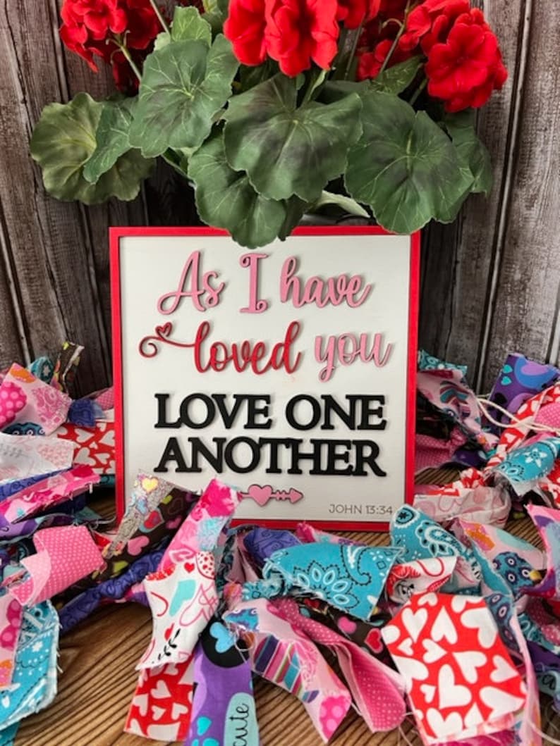 Op de afbeelding: Een wit houten bord met rode rand en de woorden "As I have loved you Love One Another John 15:34" in roze en zwarte letters. Het bord is omringd door kleurrijke stoffenresten met harten.
