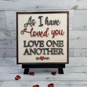 Op de afbeelding: Een houten bord met de tekst "Zoals ik van jullie heb gehouden, houd ook jullie van elkaar" en de bijbelverwijzing "Johannes 13:34". Het bord is geschilderd in grijstinten en rood en heeft een klein rood hart met een pijl.