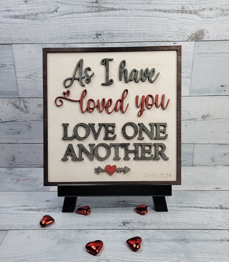 Op de afbeelding: Een houten bord met de tekst "Zoals ik van jullie heb gehouden, houd ook jullie van elkaar" en de bijbelverwijzing "Johannes 13:34". Het bord is geschilderd in grijstinten en rood en heeft een klein rood hart met een pijl.