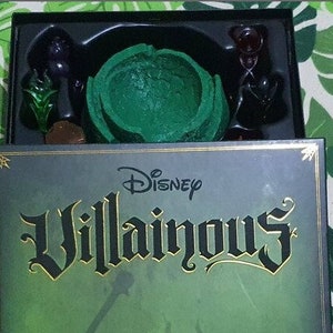 Puede incluir: Una caja del juego de mesa Disney Villainous con un diseño verde y dorado. La caja está abierta, revelando piezas del juego, incluyendo un objeto verde esculpido y figuras de personajes negros. El texto "Disney Villainous" se muestra de forma destacada.