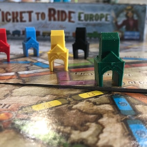 Peut inclure: Gros plan sur un plateau de jeu Ticket to Ride Europe avec six pièces de gare de différentes couleurs. Les pièces sont rouge, bleu, jaune, noir, vert et blanc.