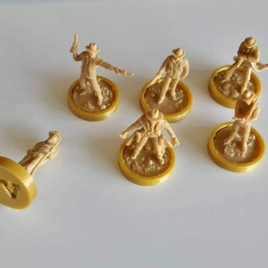 May include: Six miniature figures of cowboys, each standing on a round, gold-colored base. The bases have a textured surface and some have a number printed on them. The figures are all wearing cowboy hats and have different poses.