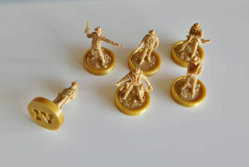 May include: Six miniature figures of cowboys, each standing on a round, gold-colored base. The bases have a textured surface and some have a number printed on them. The figures are all wearing cowboy hats and have different poses.
