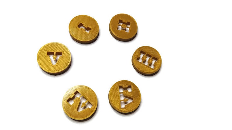 May include: Six gold-colored tokens with different letters and symbols printed on them. The tokens are arranged in a circle on a white background.