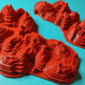 May include: Two red 3D printed models of mountains with layered peaks. The models are on a turquoise background.
