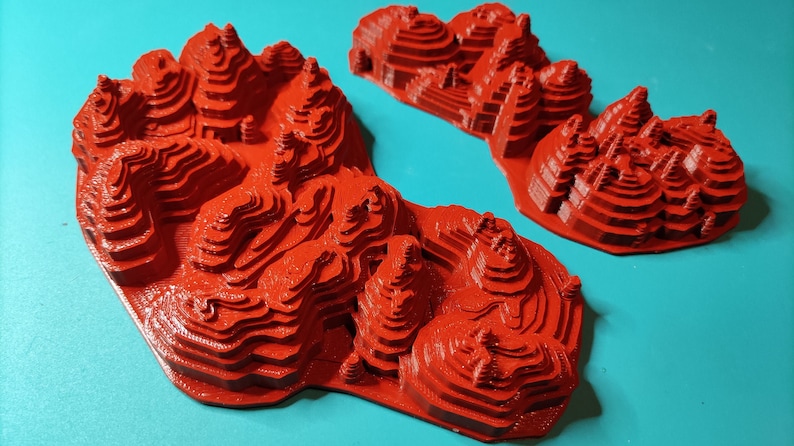 May include: Two red 3D printed models of mountains with layered peaks. The models are on a turquoise background.