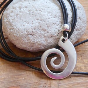 Könnte beinhalten: Eine silberne Spiralen-Anhänger-Kette mit einem schwarzen Lederband. Der Anhänger ist ein schlichtes, modernes Design mit einer glatten, polierten Oberfläche.