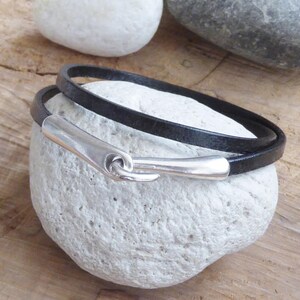 Op de afbeelding: Een zwart leren armband met een zilveren haakgesp. De armband is dubbelgevouwen en wordt getoond op een witte steen.
