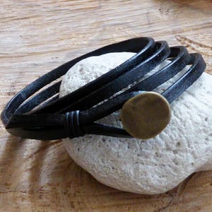 Può includere: Un braccialetto avvolgente in pelle nera con un accento rotondo color ottone. Il braccialetto è avvolto attorno a una roccia bianca.