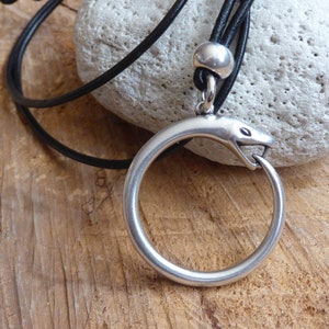 Puede incluir: Un collar con un colgante de serpiente de plata en un cordón de cuero negro. La serpiente está enroscada en un círculo con la cabeza mirando al espectador.