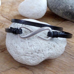 Könnte beinhalten: Ein schwarzes Lederarmband mit einem silbernen Unendlichkeits-Symbol-Anhänger. Das Armband ist verstellbar und hat einen Hakenverschluss.