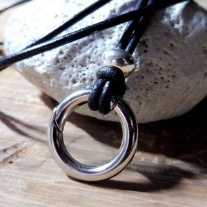 Può includere: Una collana in pelle nera con un ciondolo ad anello argentato e una perla argentata. La collana è appoggiata su una pietra bianca.